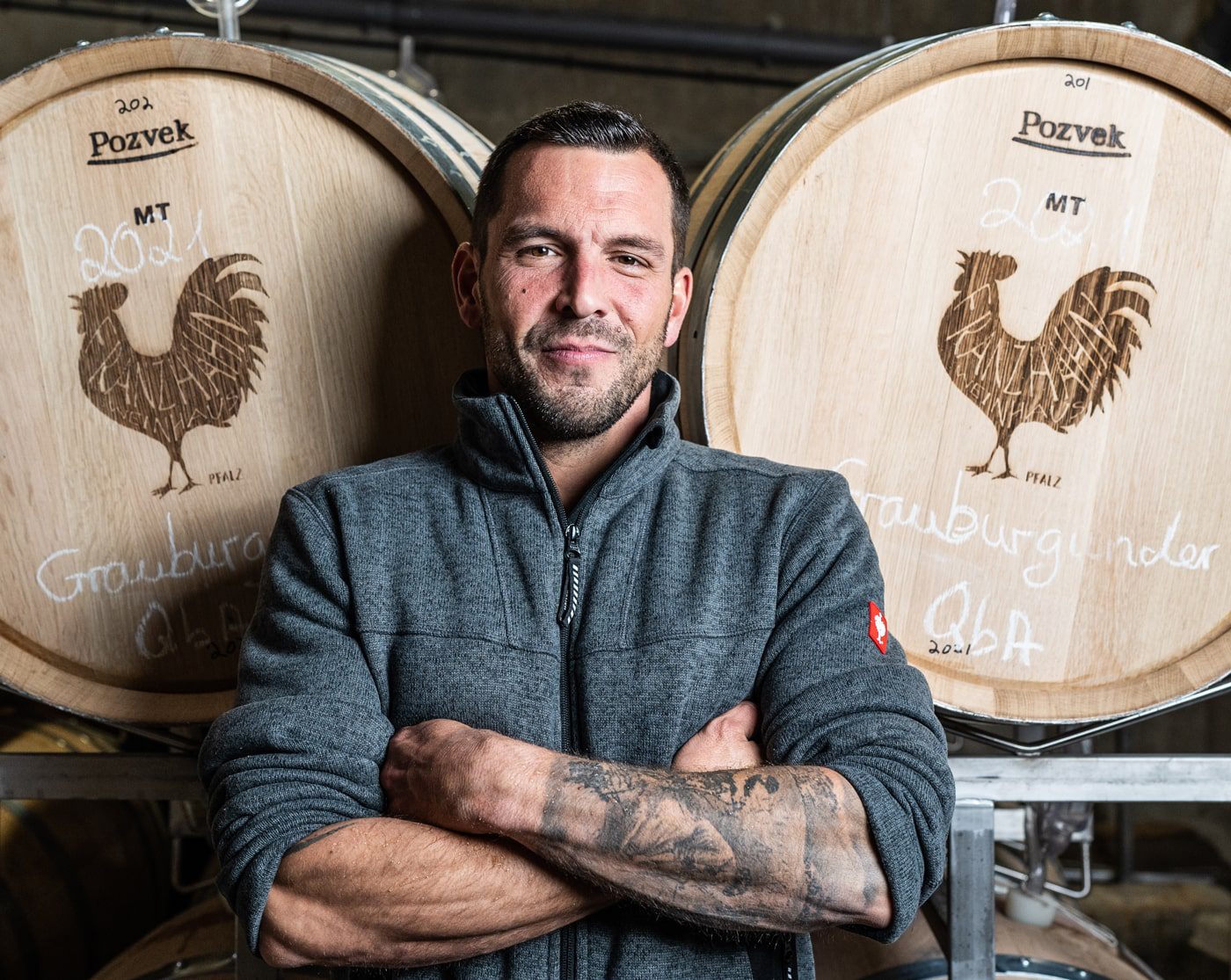 Martin Hochdörffer Ein Mann mit Tätowierungen auf den Unterarmen steht mit verschränkten Armen vor großen hölzernen Weinfässern, die jeweils mit einem Hahn und handgeschriebenen Etiketten versehen sind. Er trägt eine graue Jacke mit Reißverschluss und lächelt selbstbewusst.