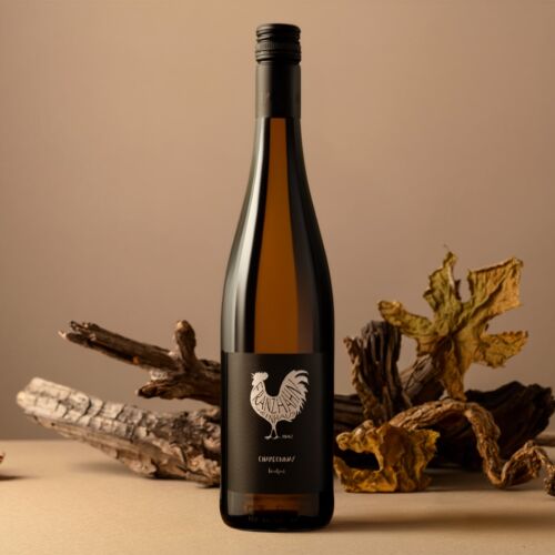 Eine Weißweinflasche mit einem schwarzen Etikett, auf dem ein weißer Hahn abgebildet ist, steht aufrecht auf einer beigen Fläche. Im Hintergrund sind getrocknete Blätter und Holzstücke dekorativ angeordnet.