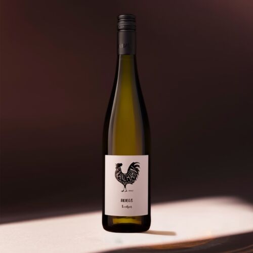 Eine Weinflasche mit einem schwarzen Schraubverschluss steht auf einer sonnenbeschienenen Fläche. Das Etikett ist weiß mit einem schwarzen Hahn und darunter liegendem Text, der vor einem dunklen, leicht verschwommenen Hintergrund steht.