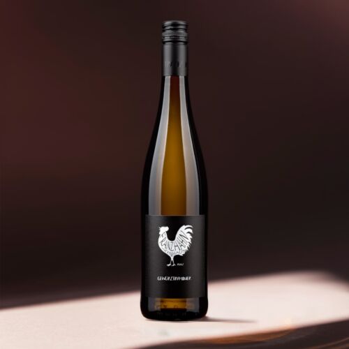 Eine hohe, dunkle Weinflasche mit einem schwarzen Etikett, auf dem ein weißer Hahn und das Wort "Gewürztraminer" abgebildet sind, steht auf einer sanft beleuchteten Fläche vor einem verschwommenen, dunkelbraunen Hintergrund.