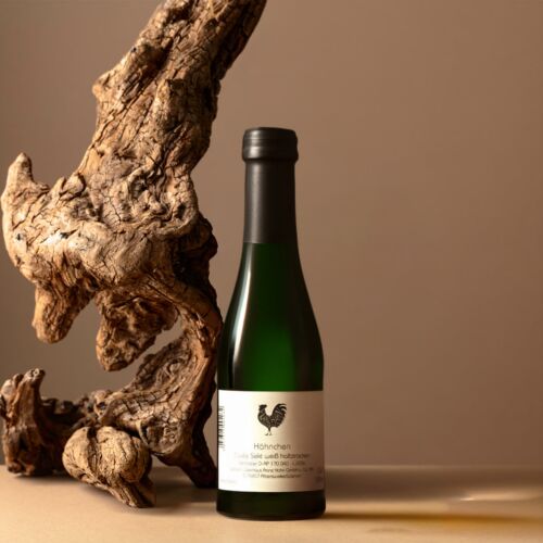 Eine kleine grüne Weinflasche mit der Aufschrift "Hähnchen" und einem schwarzen Verschluss steht auf einer beigen Fläche neben einem großen, strukturierten Stück Treibholz vor einem neutralen Hintergrund.