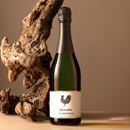 Eine Sektflasche mit einem weißen Etikett, auf dem ein schwarzer Hahn abgebildet ist, steht auf einer beigen Fläche neben einem großen, strukturierten Stück Treibholz vor einem neutralen Hintergrund.