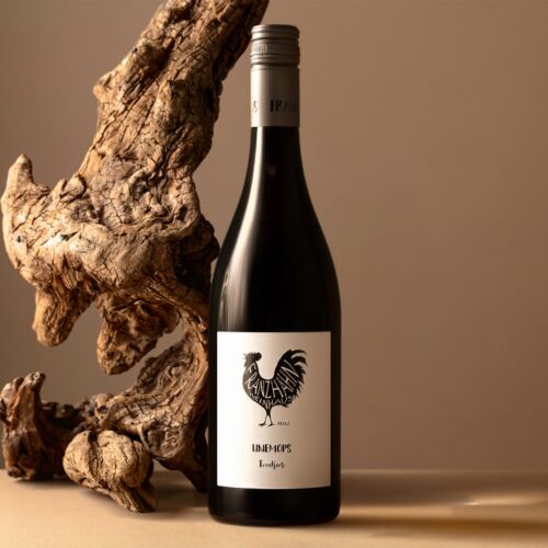 Eine Weinflasche mit einem weißen Etikett, auf dem ein schwarzer Hahn abgebildet ist, steht auf einer beigen Fläche neben einem strukturierten Stück Treibholz, vor einem neutralen braunen Hintergrund.
