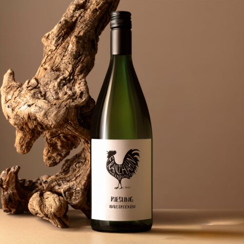 Eine Flasche Rieslingwein mit einem schwarzen Hahn auf dem Etikett steht auf einer beigen Fläche neben einem großen, strukturierten Stück Treibholz vor einem neutralen Hintergrund.