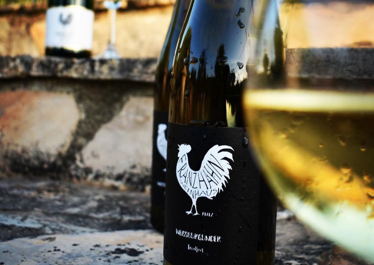 Nahaufnahme einer Weinflasche mit schwarzem Etikett und weißem Hahn-Logo, daneben ein Glas Weißwein. Andere Flaschen und Steinstufen sind im Hintergrund unscharf.