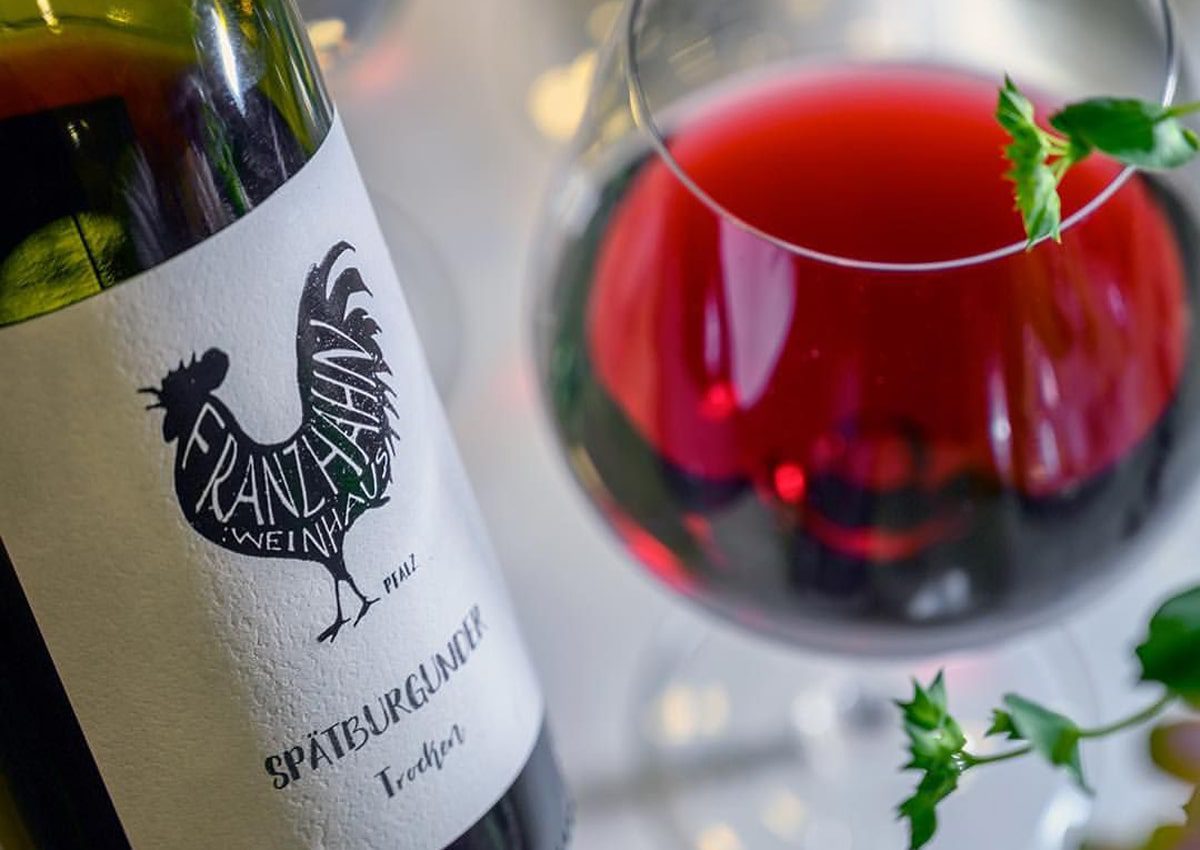 Nahaufnahme einer Weinflasche mit der Aufschrift "Spätburgunder Trocken" und einem Hahn neben einem mit Rotwein gefüllten Glas, daneben eine grüne Blattgarnitur.