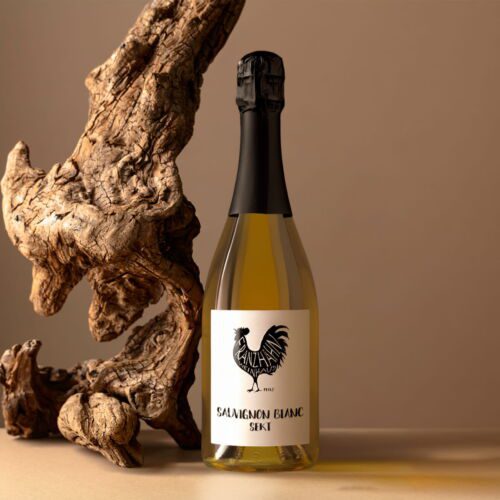 Eine Flasche Sauvignon Blanc Sekt mit einem Hahn-Logo auf dem Etikett steht neben einem verdrehten Stück Treibholz auf einer beigen Fläche und Hintergrund.