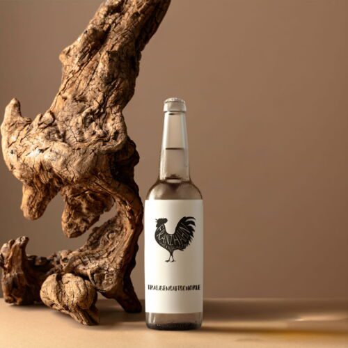 Eine Flasche Traubensaftschorle mit dem Logo eines Hahns und "TRALE&AMSHCIPPLE" auf dem Etikett steht neben einem großen Stück Treibholz auf einem neutralen Hintergrund.