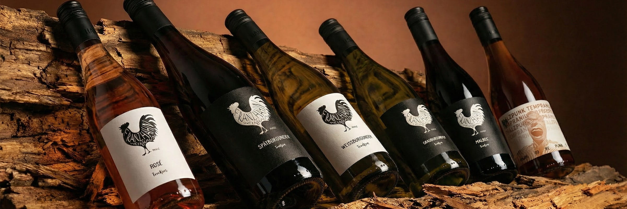 Sechs Weinflaschen mit schwarzen Hahn-Etiketten sind auf rauen Holzstämmen ausgestellt. Bei den Flaschen handelt es sich um Rosé-, Weiß- und Rotweine, die schräg vor einem warmen, braunen Hintergrund stehen.
