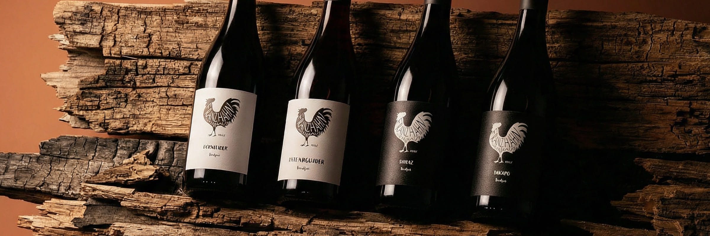 Vier Weinflaschen mit Hahnmotiven auf den Etiketten sind in einer Reihe auf rustikalen Holzstämmen vor einem warmen braunen Hintergrund angeordnet.