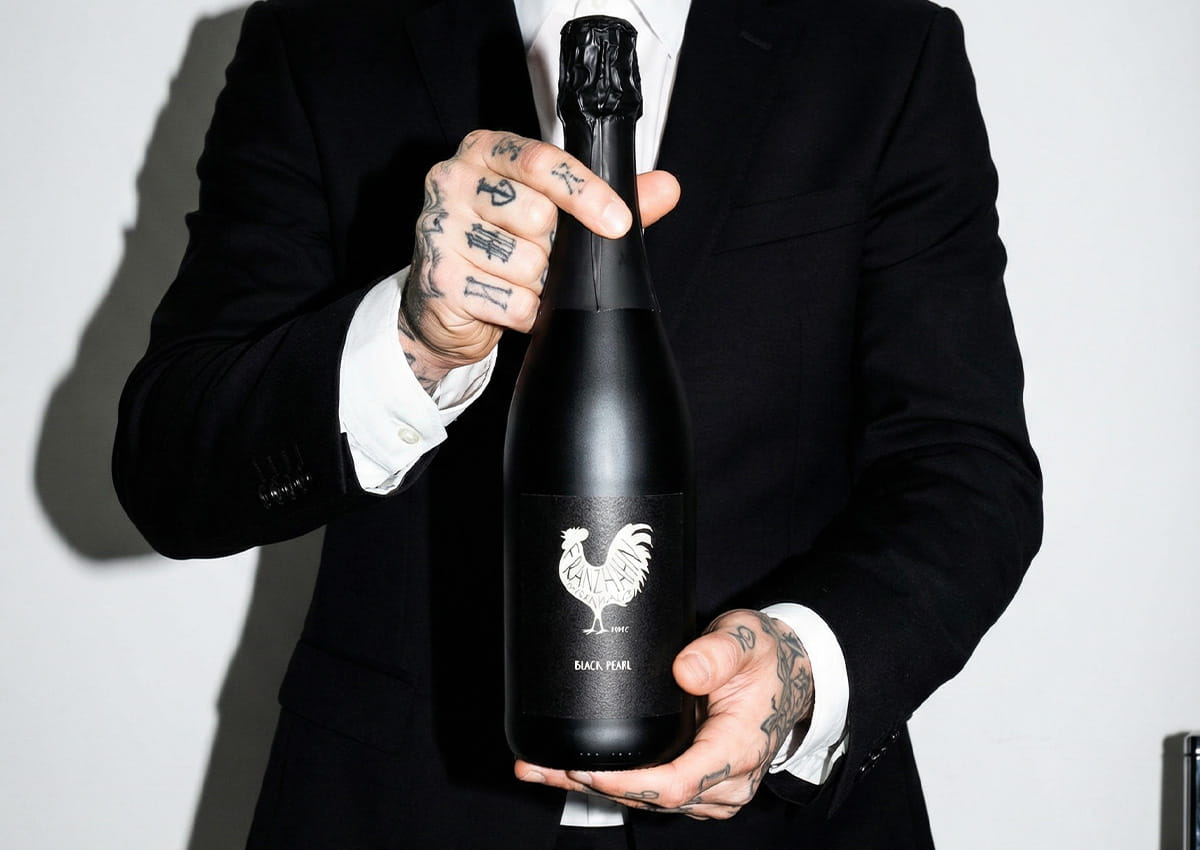 Eine Person in einem schwarzen Anzug mit tätowierten Händen hält eine schwarze Sektflasche mit einem weißen Hahn-Logo auf dem Etikett vor einem schlichten Hintergrund.